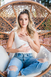 A woman in blue jeans and a white top sits in a chair, smiling, with her hands gently placed on her chest.